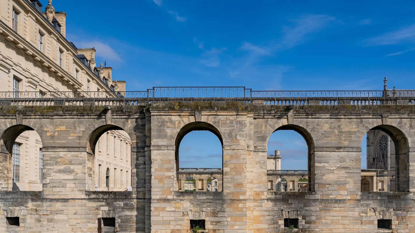 Masmavi gökyüzü altında Château de Vincennes'in içindeki kemerli taş duvarlar, uzaktaki kemerde Sainte-Chapelle'in gül penceresi çerçevelenmiş